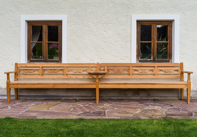 Geschnitzte Holzbank vor ländlicher Hausfassade mit zwei Holzfenstern und Steinpflaster.