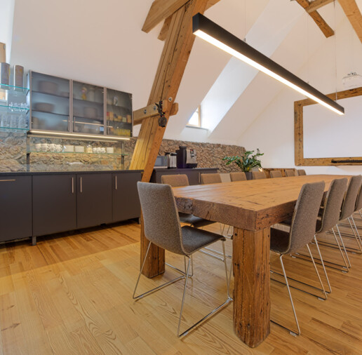 Besprechungsraum mit Möbeln aus Altholz: langer Konferenztisch mit rustikalen Balken, moderne Hängeleuchte, Küchenzeile und Leinwand.