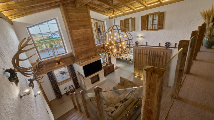 Großer Wohnraum im Chaletstil mit Altholz-Decke, sichtbaren Balken, Glasgeländer, Kugelleuchte, Kamin und Schiebetür – Innenausbau aus Altholz.