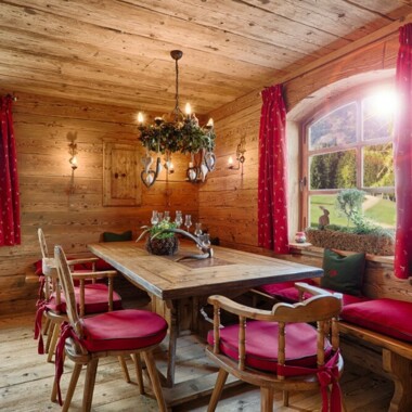 Stube mit Esstisch, Bänken und Stühlen aus Altholz, roten Vorhängen und Bergblick.