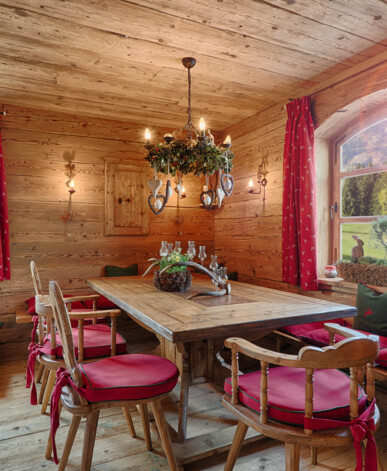 Gemütliche Stube mit Möbeln aus Altholz: großer Esstisch, Bank und Stühle mit roten Polstern, Bergblick durchs Fenster, schlafender Hund.