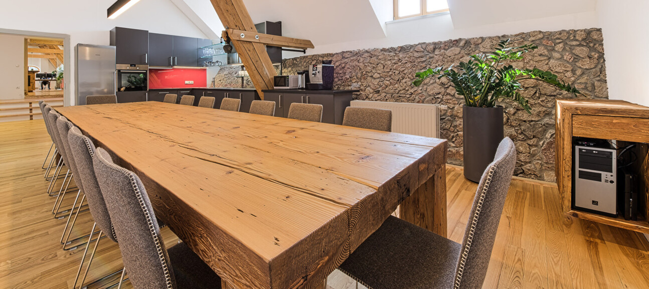 Ein moderner Raum mit langem Esstisch aus Altholz, sichtbaren Dachbalken und einer Küchenzeile.