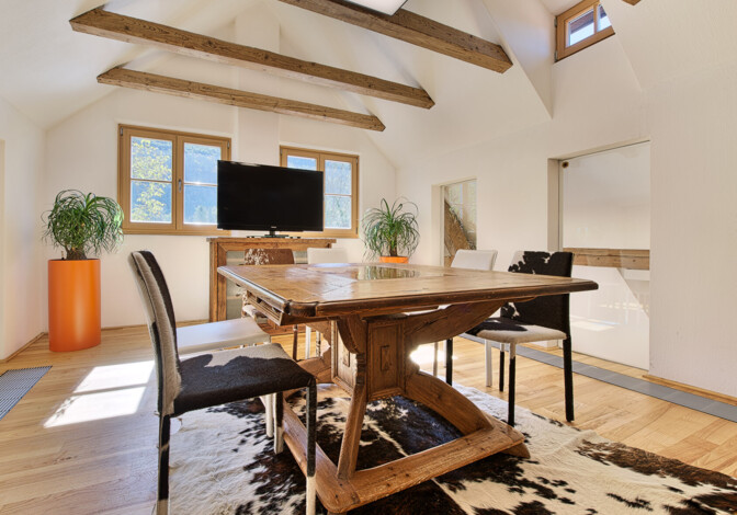 Heller Dachraum mit handgefertigtem Altholz-Tisch, sichtbaren Deckenbalken und Kuhfellteppich.