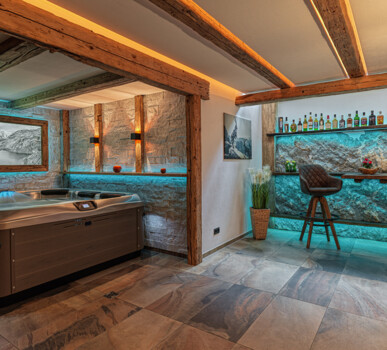 Wellnessraum mit Whirlpool, beleuchteter Bar, Deckenbalken und Möbel aus Altholz, kombiniert mit Stein- und LED-Akzenten.
