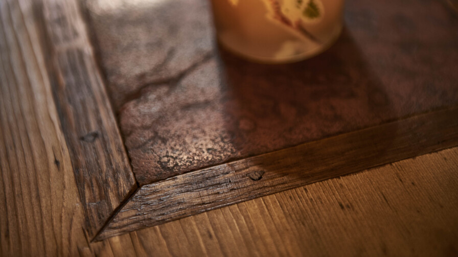 Detail einer Altholz-Tischplatte, stimmig beleuchtet von einer Kerze im Glas mit Blattdekor.