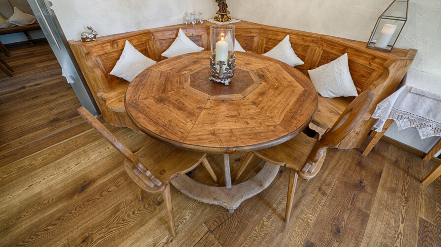 Eckbank mit rundem Tisch aus Altholz; weiße Kissen und Kerze schaffen eine gemütliche Stube.