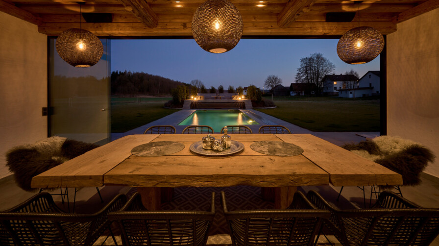 Ein stimmungsvoll beleuchteter Essbereich mit Altholz-Esstisch unter Sichtbalken bietet Blick auf den beleuchteten Pool bei Abenddämmerung.