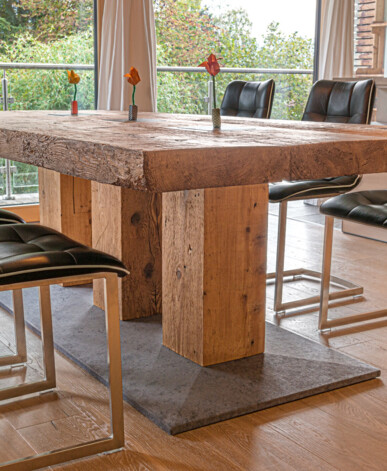 Esstisch aus Altholz mit schwarzen Lederstühlen in einer hellen Wohnküche mit Blick ins Grüne.