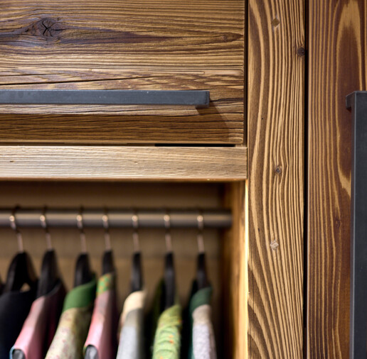 Nahaufnahme eines Altholz-Kleiderschranks mit Metallgriffen und Kleiderstange mit hängender Kleidung.