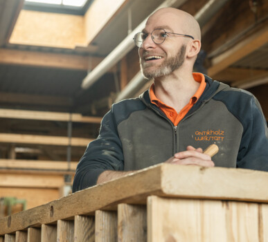 Lächelnder Tischler in der Antikholzwerkstatt an der Werkbank, mit Holzstück in der Hand, umgeben von Balken und Altholz.