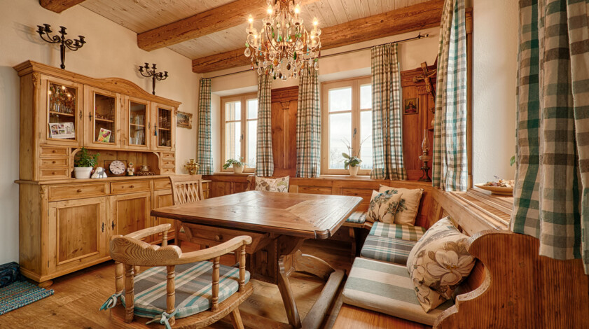 Eine gemütliche Bauernstube aus Altholz mit Eckbank, Tisch und Kredenz, holzgetäfelten Wänden, karierten Vorhängen und Kronleuchter.
