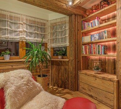 Gemütliche Leseecke mit Altholz-Wandverkleidung und beleuchtetem Bücherregal. Im Vordergrund steht ein roter Sessel mit Fell.
