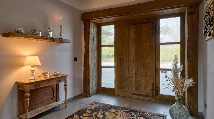 Eingangsbereich mit Altholz-Haustür, seitlichen Fensterfeldern, Konsole mit Lampe und Teppich.