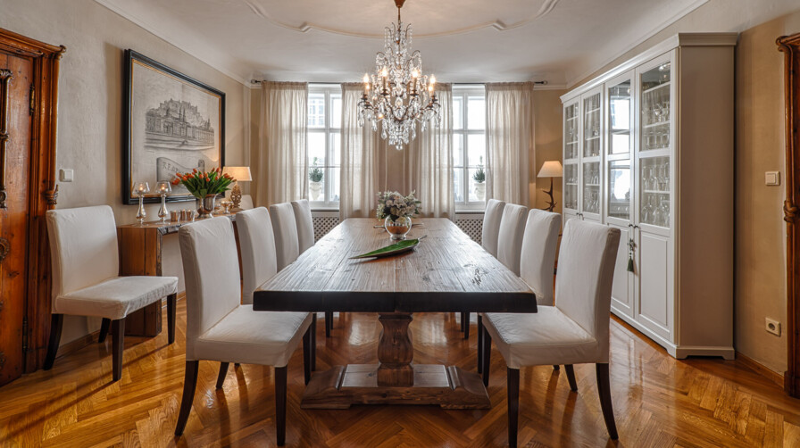 Elegantes Esszimmer mit Esstisch aus Altholz, weißen Polsterstühlen, Kristallkronleuchter und Glasvitrine auf Fischgrätparkett.