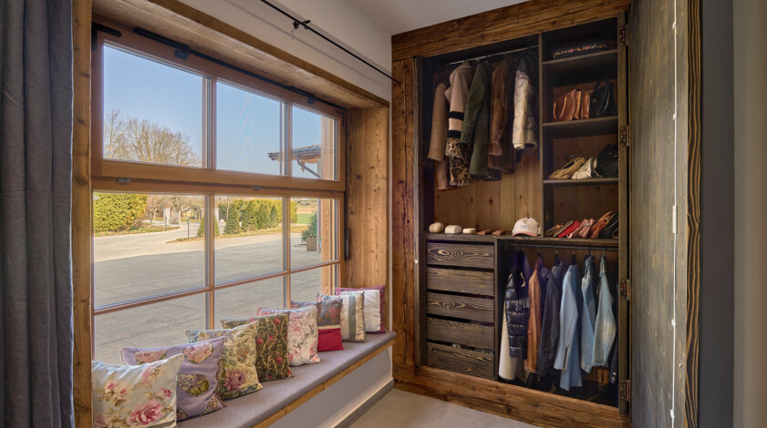 Offener Garderobenschrank aus Altholz neben Sitzfenster mit bunten Kissen und Blick in den Garten.