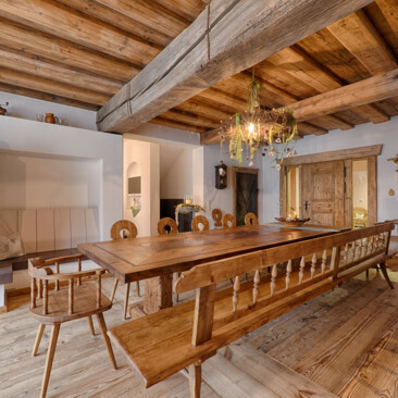 Essbereich mit Möbeln aus Altholz: langer Tisch, Bank und Stühle unter Deckenbalken, neben Kamin und Holztür.
