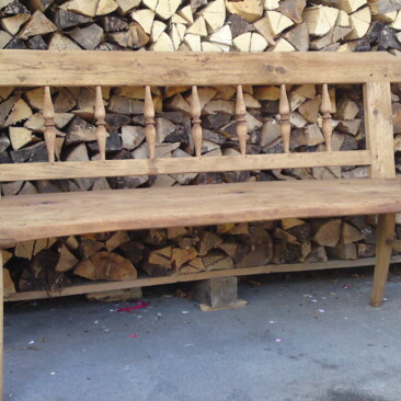 Bank aus Altholz, mit gedrechselter Rückenlehne vor gestapeltem Brennholz.