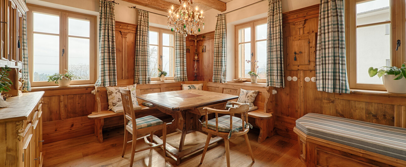 Gemütliche Bauernstube aus Altholz mit Sichtbalken, Eckbank, Holztisch, karierten Vorhängen und Kronleuchter.