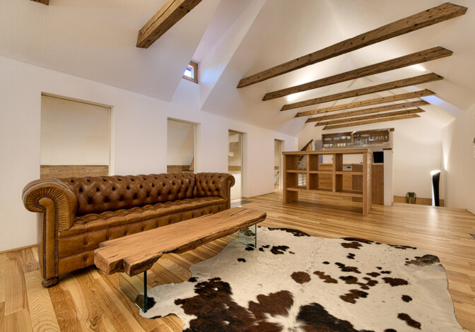 Helles Dachstudio mit Ledersofa, Kuhfellteppich und Couchtisch aus Altholz. An der Decke sichtbare Balken und Möbel aus Altholz in Küche und Wohnbereich.
