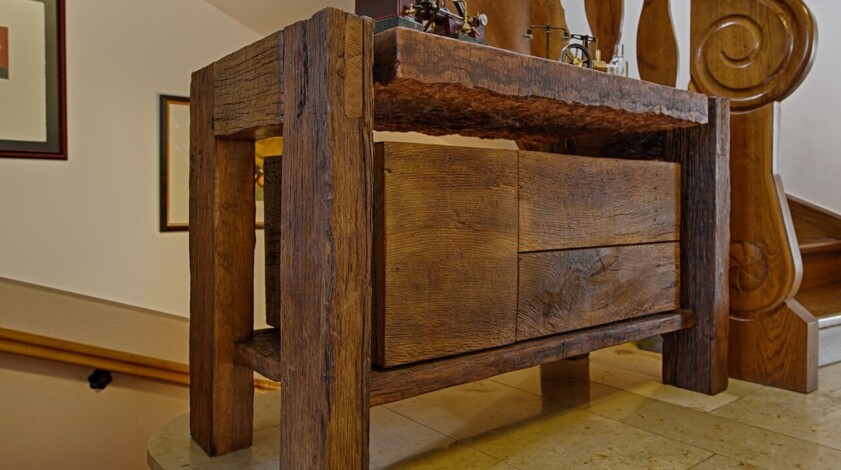 Rustikale Konsole aus Altholz mit Schubladen, auf einem Treppenabsatz neben geschnitztem Holzgeländer.
