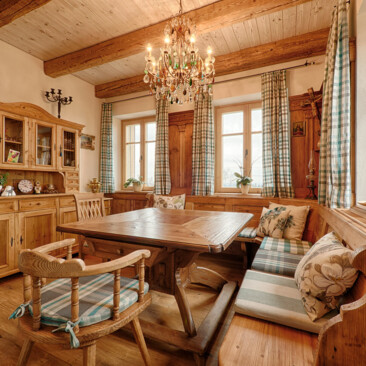 Eine gemütliche Bauernstube aus Altholz mit Eckbank, Tisch und Kredenz, holzgetäfelten Wänden, karierten Vorhängen und Kronleuchter.