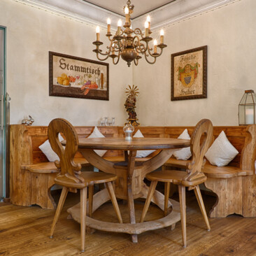 Gemütliche Stube mit runder Eckbank, Tisch und Stühlen, handgefertigten Möbeln aus Altholz, Kronleuchter und Stammtisch-Wandbild.