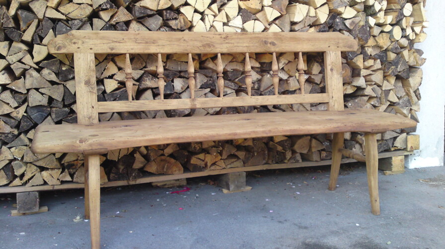 Bank aus Altholz, mit gedrechselter Rückenlehne vor gestapeltem Brennholz.