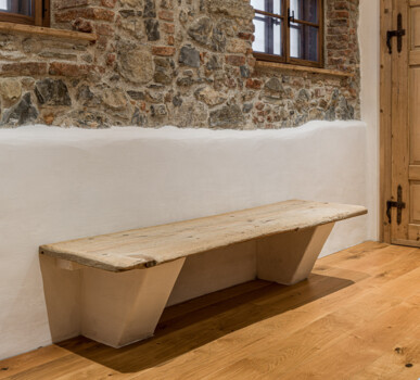 Rustikaler Flur mit Bruchsteinwand, Holzfenstern und massiver Altholz-Tür; davor eine schlichte Sitzbank auf Holzboden.