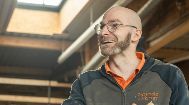 Lächelnder Tischler in der Antikholzwerkstatt an der Werkbank, mit Holzstück in der Hand, umgeben von Balken und Altholz.