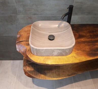 Ein quadratisches Steinwaschbecken steht auf einer massiven Altholzplatte mit Naturkante und schwarzer Armatur im modern-rustikalen Stil.