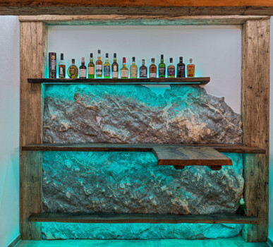 Maßgefertigtes Barregal aus Altholz vor Naturstein, mit türkisfarbener LED-Beleuchtung und einer Reihe Spirituosenflaschen.