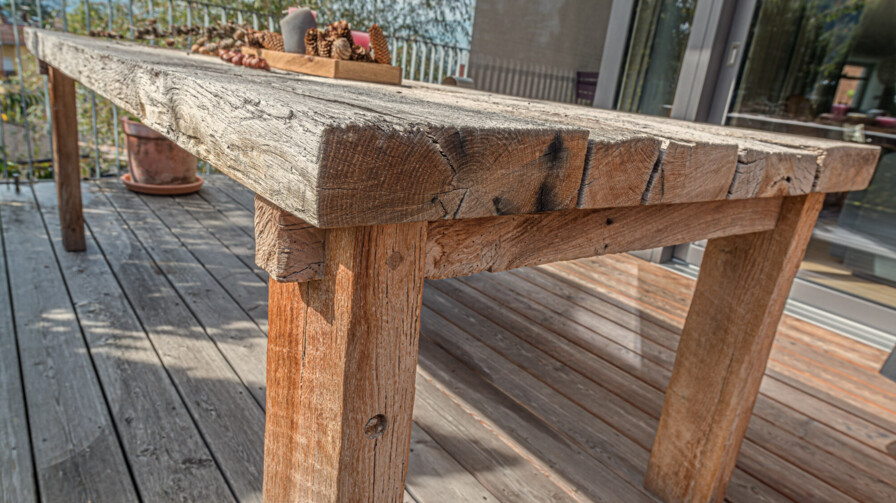 Massiver Tisch aus Altholz auf einer Terrasse, mit rustikaler, rissiger Tischplatte und naturbelassener Patina.