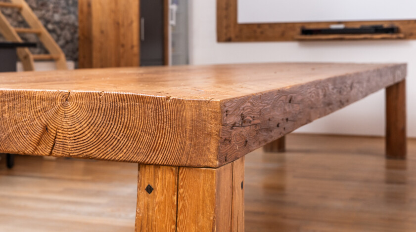 Nahaufnahme eines massiven Tisches aus Altholz mit sichtbarer Jahresring- und Balkenstruktur in einem modernen Raum.