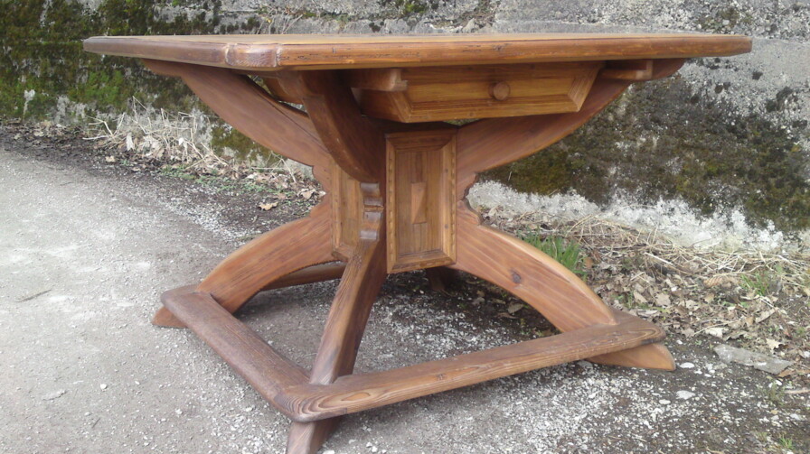 Holztisch mit Kreuzgestell und zentralem Kasten aus Altholz, im Freien vor einer Steinmauer.