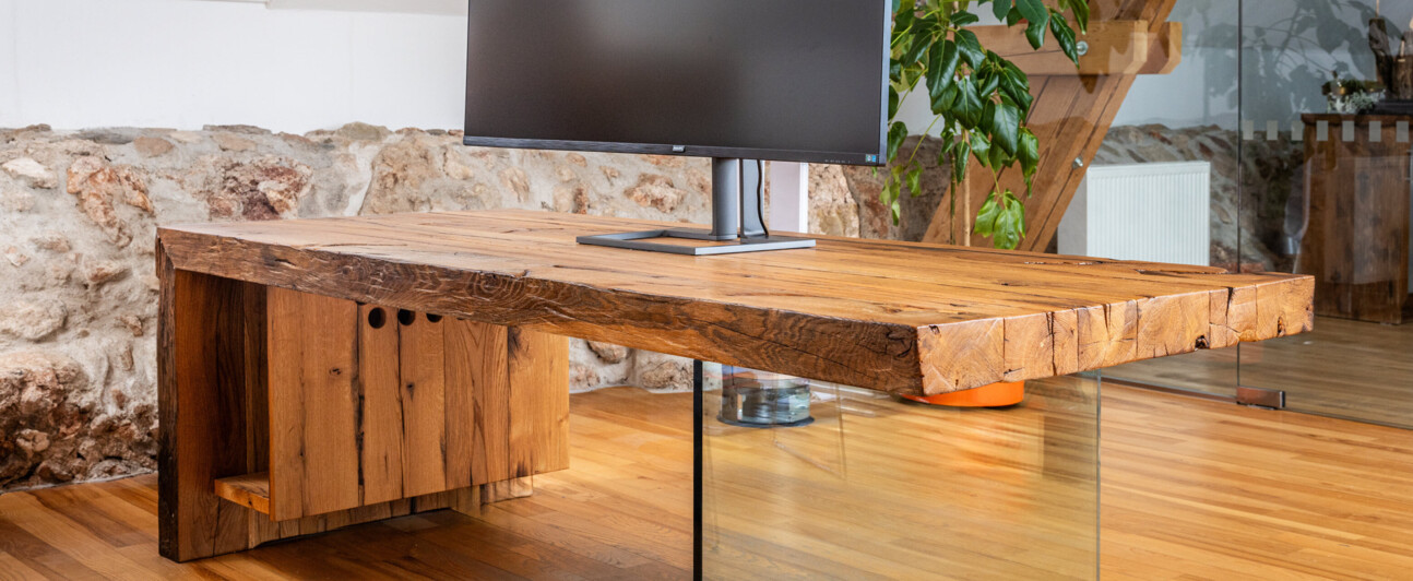 Schreibtisch aus Altholz mit Glasfuß und Curved-Monitor, in einem hellen Loftbüro.