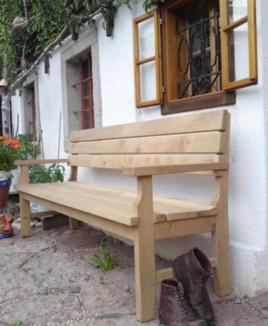 Holzbank aus Altholz vor weißer Hausfassade mit geöffnetem Holzfenster; daneben stehen abgetragene Lederstiefel.