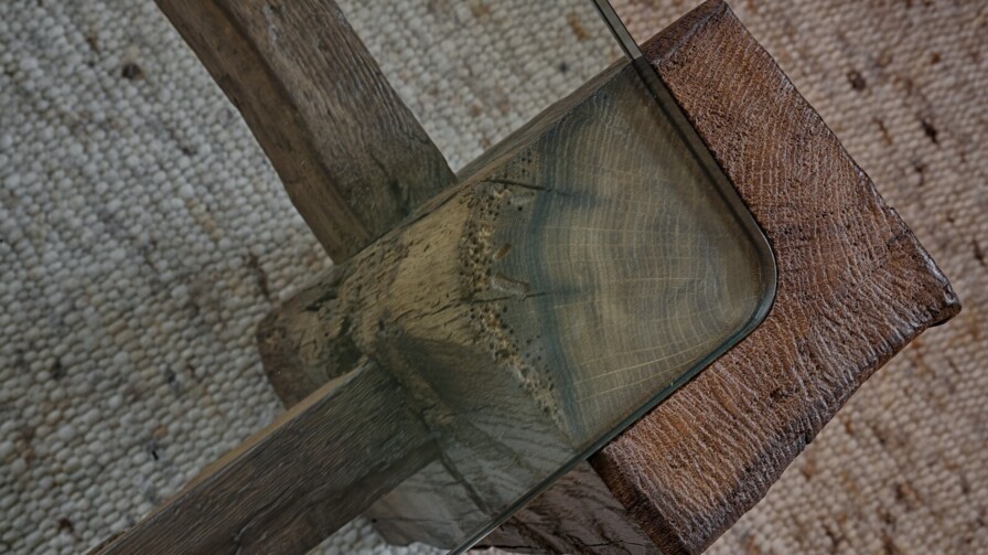 Detail eines Tisches mit Glasplatte auf massivem Altholzgestell, Nahaufnahme der Ecke auf Naturteppich.