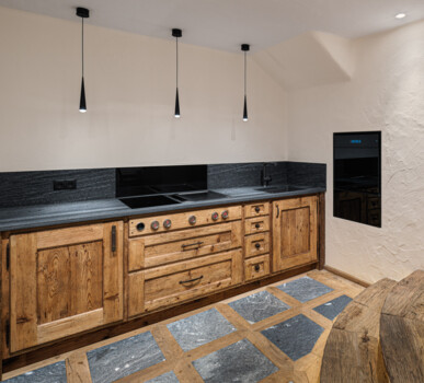 Maßgefertigte Küche aus Altholz mit schwarzer Steinplatte, Induktionskochfeld und Einbaugerät, stimmig beleuchtet neben rustikaler Tür und Stufen.