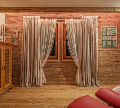Gemütliche Stube mit Fenster, Vorhängen und Sideboard vor Altholz-Wand.