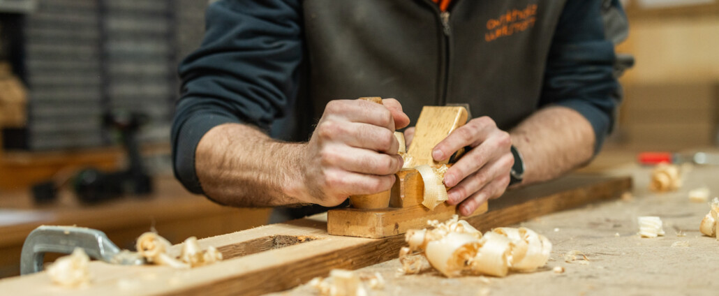 Ein Handwerker hobelt Holz von Hand, dicke Späne bedecken die Werkbank in der Antikholzwerkstatt.