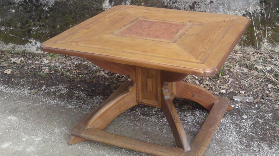 Ein quadratischer, handgefertigter Tisch aus Altholz, mit Intarsie, vor einer Steinmauer.