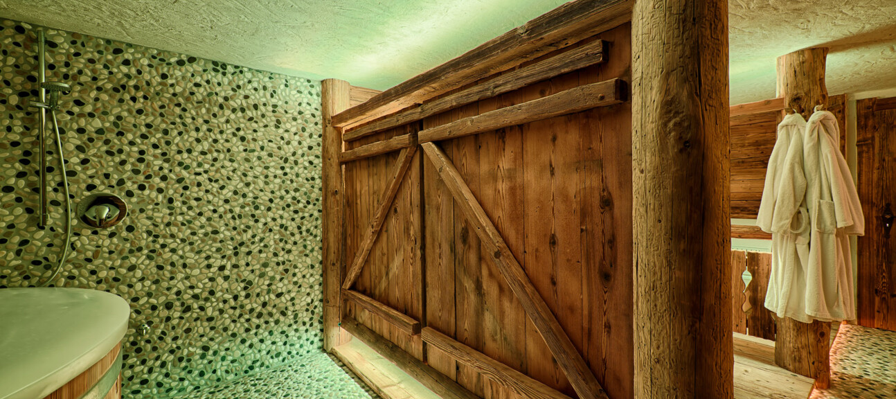 Rustikaler Wellnessbereich mit Dusche, Kieselmosaik und Möbeln aus Altholz; warme Beleuchtung und Bademäntel am Holzbalken.