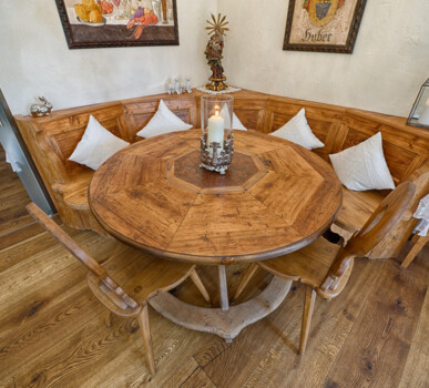 Eckbank mit rundem Tisch aus Altholz; weiße Kissen und Kerze schaffen eine gemütliche Stube.