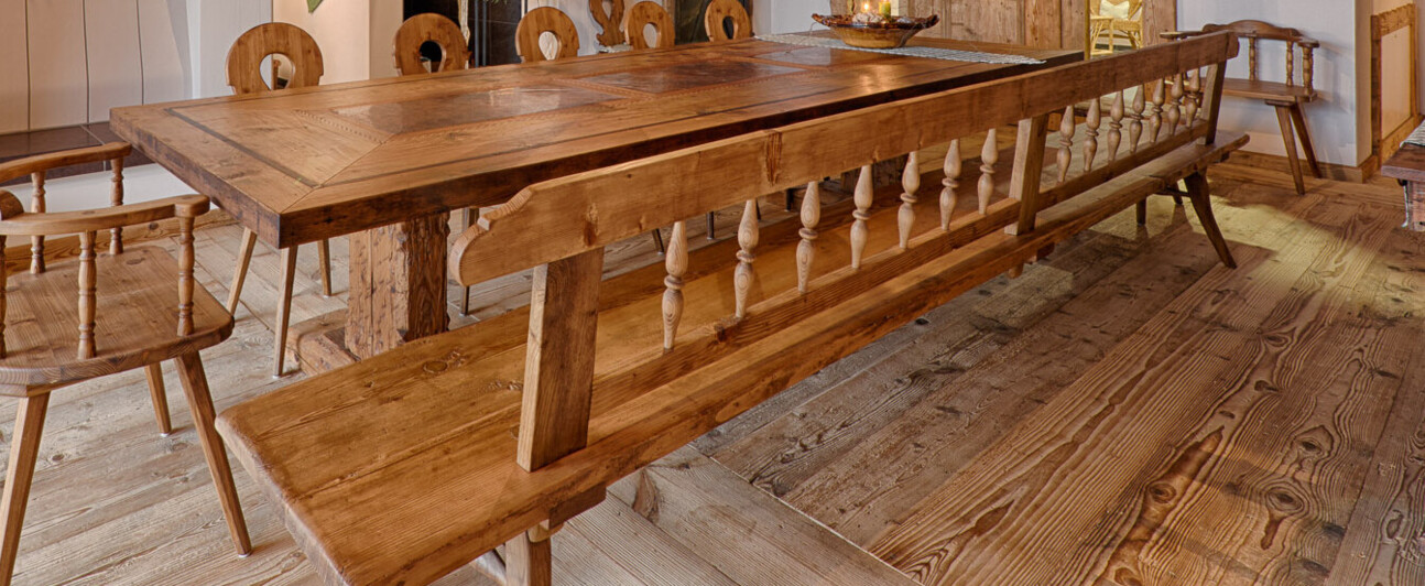 Gemütliches Esszimmer aus Altholz: Tafel mit Bank und Stühlen, sichtbare Deckenbalken, Holzboden, Kamin und naturhafte Leuchte.