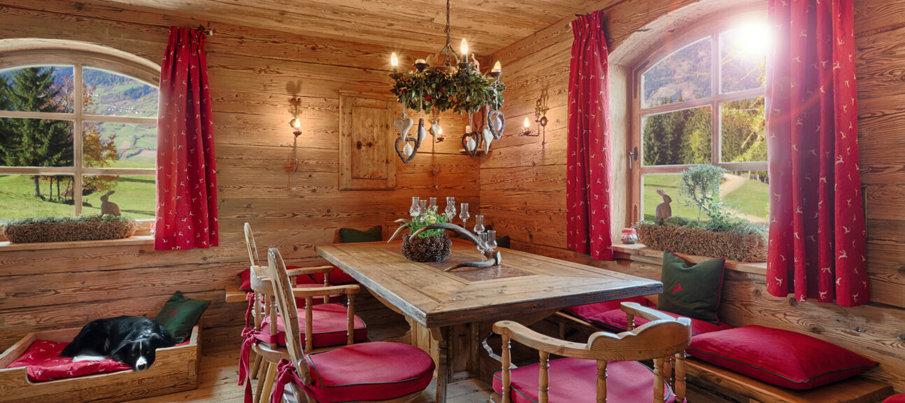 Gemütliche Stube mit Möbeln aus Altholz: großer Esstisch, Bank und Stühle mit roten Polstern, Bergblick durchs Fenster, schlafender Hund.