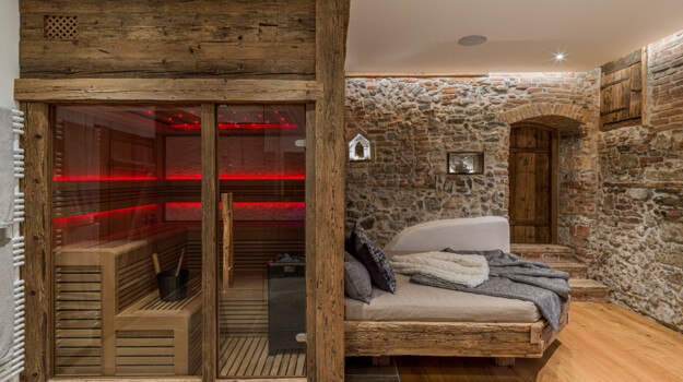 Wellnessraum mit Sauna aus Altholz, rot beleuchtet, daneben ein Bett auf Altholzrahmen vor einer Natursteinwand.