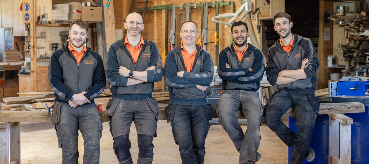 Fünf Tischler der Antikholzwerkstatt posieren in der Werkstatt, in Arbeitskleidung zwischen Werkbänken, Maschinen und Holz.