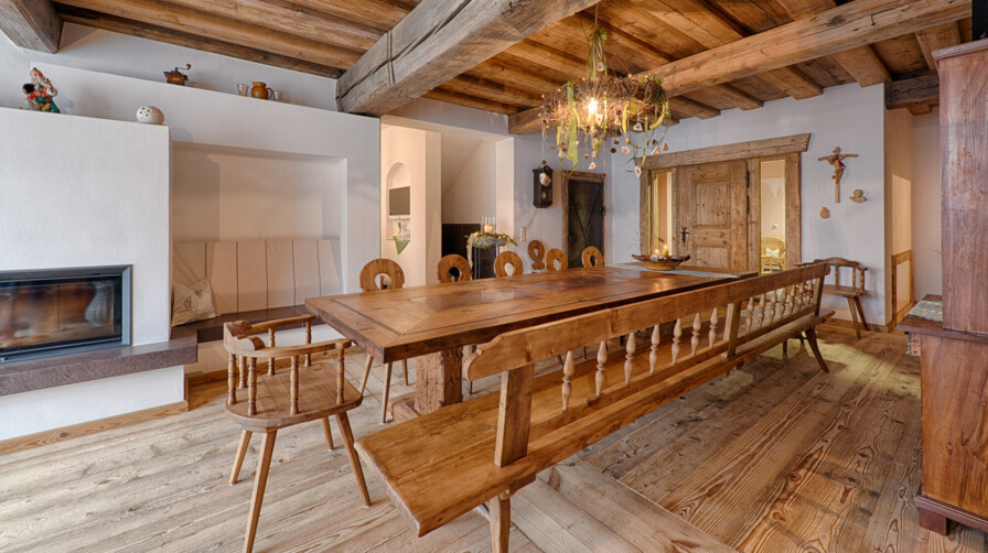 Essbereich mit Möbeln aus Altholz: langer Tisch, Bank und Stühle unter Deckenbalken, neben Kamin und Holztür.