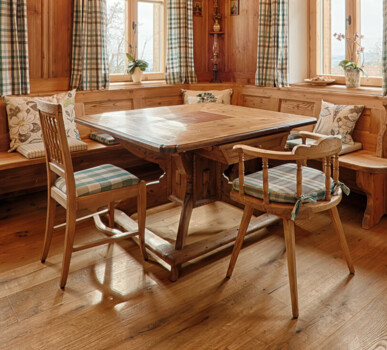 Gemütliche Bauernstube aus Altholz mit Sichtbalken, Eckbank, Holztisch, karierten Vorhängen und Kronleuchter.