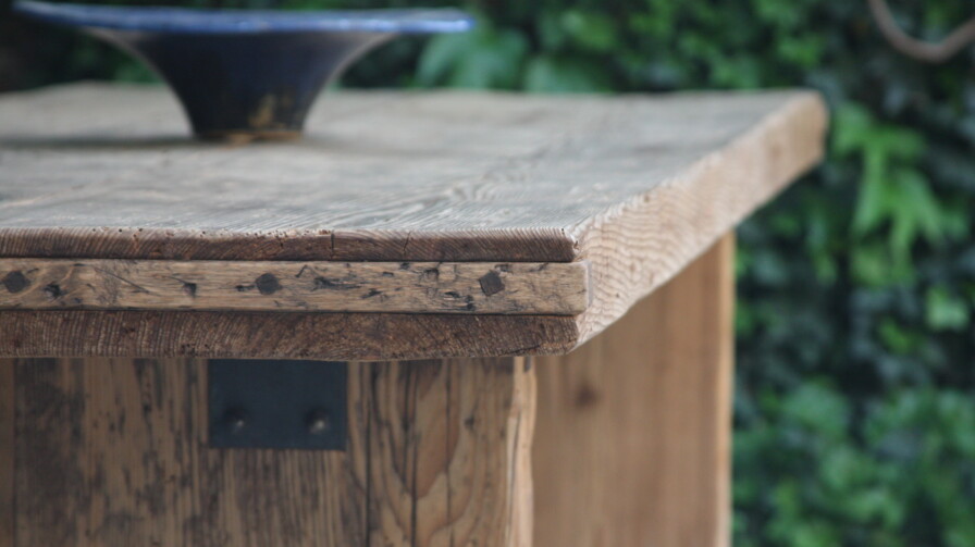 Detail eines handgefertigten Altholz-Tisches mit rustikaler Kante; oben eine dunkle Keramikschale, dahinter grünes Laub.